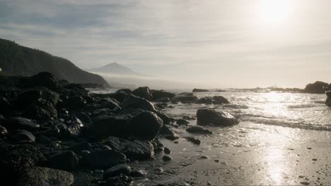 Etérea-Playa-De-Arena-Negra-Al-Atardecer-Con-El-Pico-Del-Teide-A-Distancia,-Rocas-Brumosas-Con-Suaves-Olas-Al-Amanecer,-Tenerife-Islas-Canarias