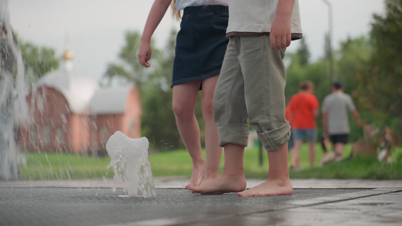 Barefoot teenagers using leg to touch water fountain jet on metal grate, neighborhood houses and people in background, summer park scene showing playful curiosity and urban leisure