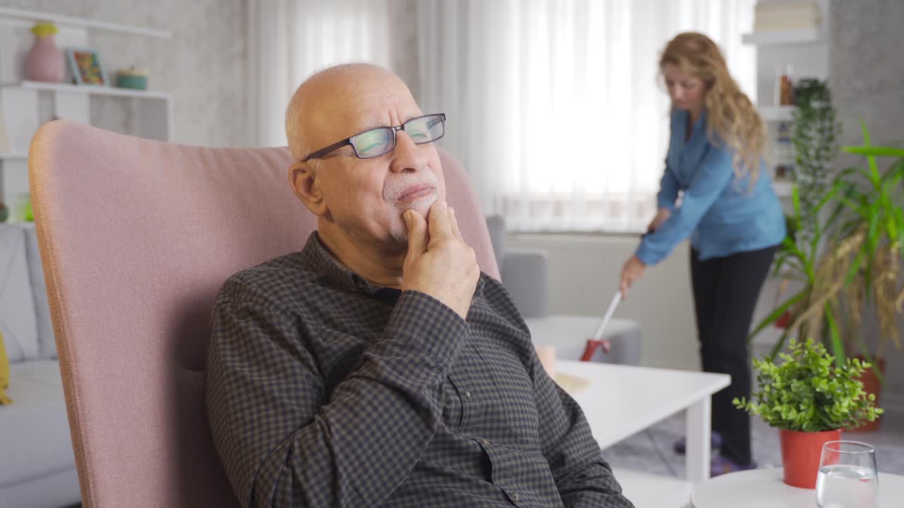 The old man is thoughtful and in the background his wife is cleaning.