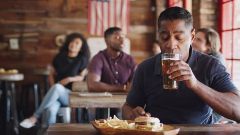 mature man watching game on screen in sports bar eating burger and fries and drinking beer