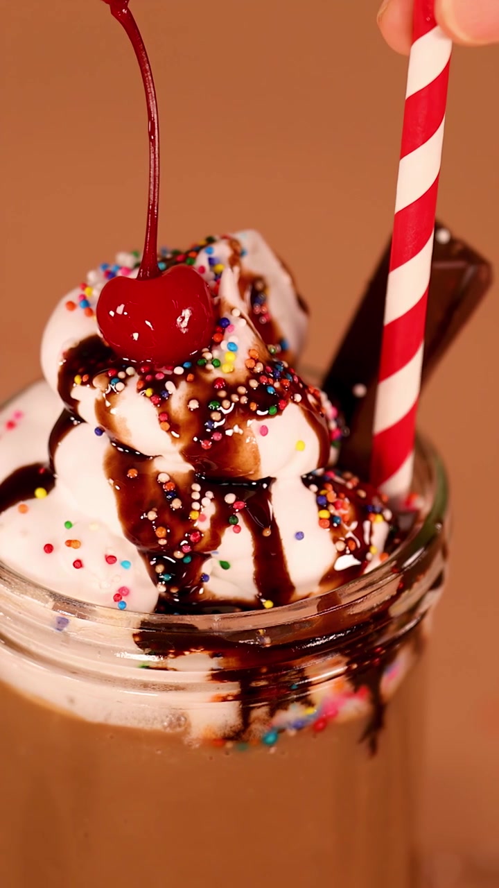 Primo piano di un cremoso milkshake al cioccolato con panna montata mentre una mano inserisce una cannuccia a strisce, illuminazione calda