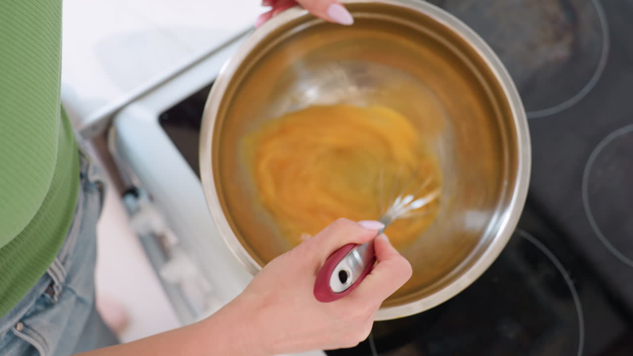 Top down view of chef whisking egg mixture in stainless steel bowl, blending yolk and white together during baking preparation process, handheld kitchen utensil mixing technique