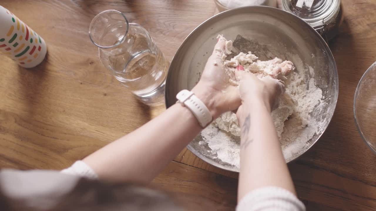 Overhead shot of a woman kneading fresh dough in a metal bowl on a rustic wooden kitchen table during baking preparation