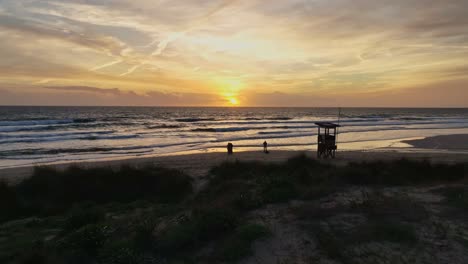 Sillouettes-of-people-walking-along-the-beach-at-high-tide-with-sun-dropping-below-horizon