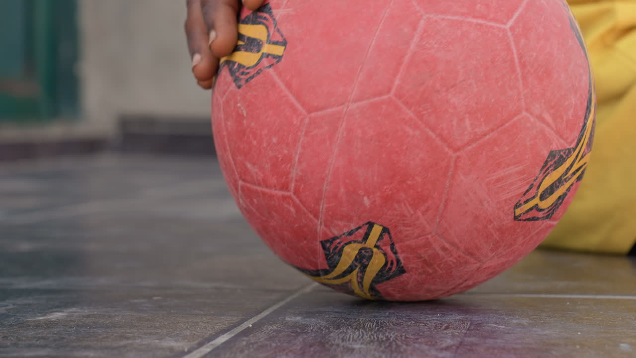 Palla da calcio rossa inquadrata dal basso, mano nera che la fa rotolare su piastrelle, dita di bambino nero che ne guidano il movimento vicino a vestiti gialli, gioco spontaneo ed energico pieno di vitalità e un'atmosfera di quartiere piena di speranza, con un tocco cinematografico
