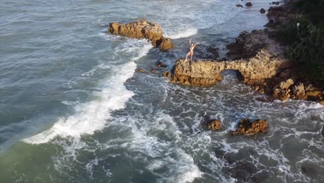 Mädchen,-Das-Auf-Einem-Felsen-Am-Rande-Des-Brasilianischen-Ozeans-Steht,-Schwenkt-Zur-Sonnenuntergangsdrohnenantenne