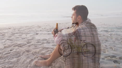 sitting on sandy beach, man wrapped in blanket holding beverage bottle
