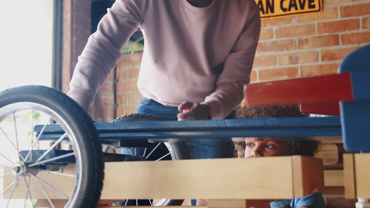 Close up of middle aged black father and his pre teen son standing at a workbench in their garage building a racing kart together, low angle