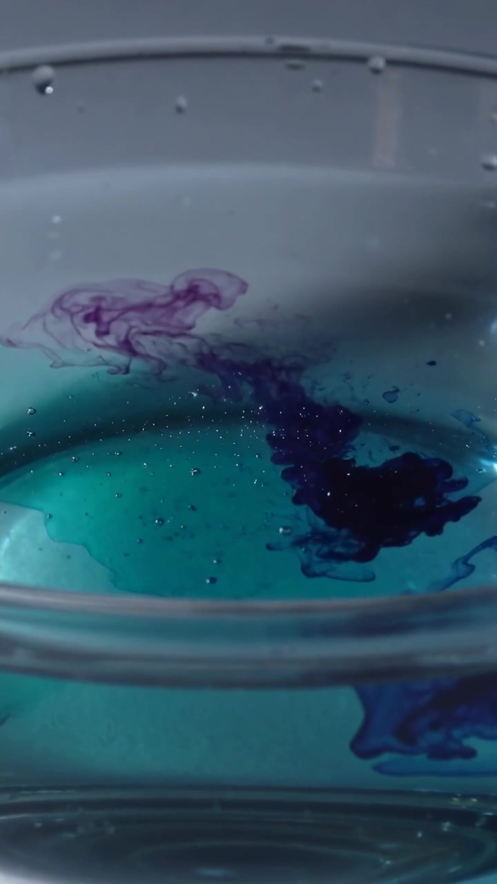 Vertical video: Dropping purple dye plume sinking, spreading in clear bowl, mixing with teal liquid