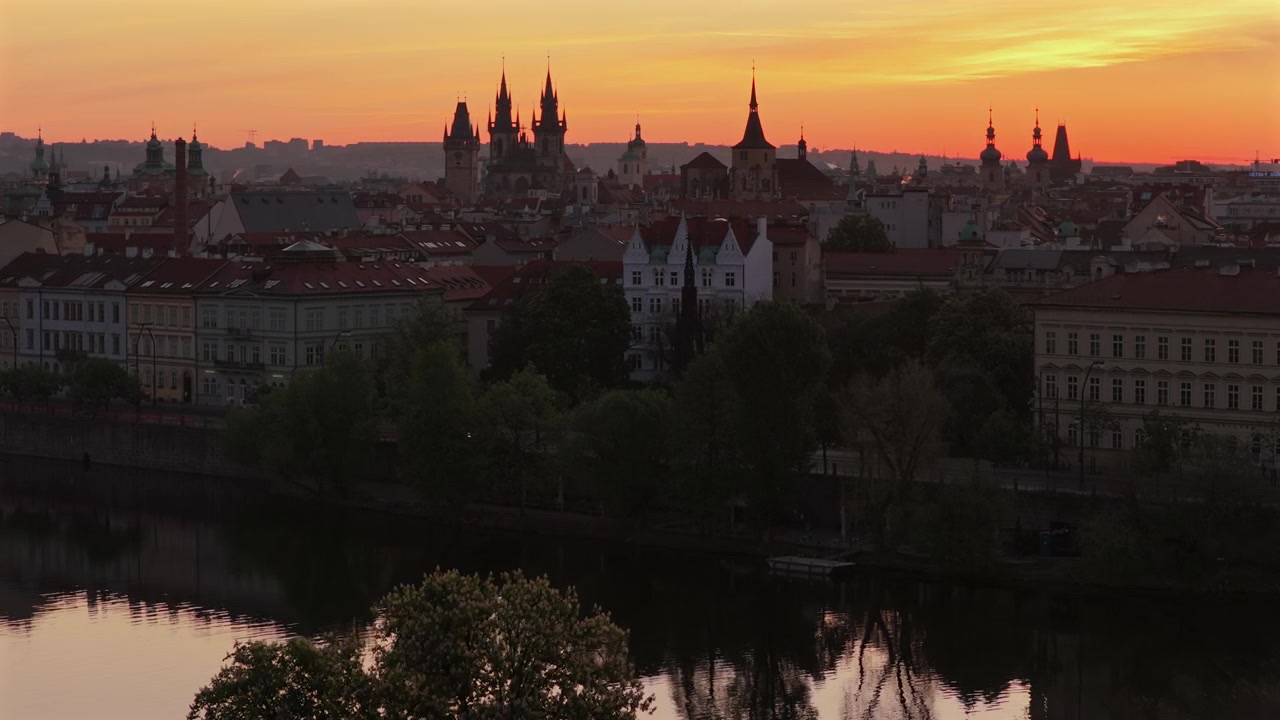 Veduta aerea con drone all'alba rosso-arancio dello skyline della Città Vecchia di Praga con le guglie della Chiesa di Týn e molteplici torri gotiche in silhouette, alberi e la riva del fiume Moldava in primo piano, Praga.