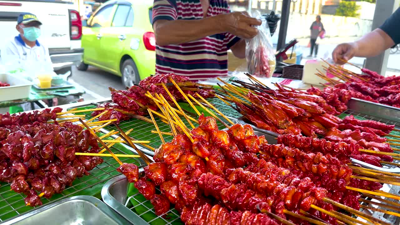 A street vendor prepares vibrant chicken skewers in a bustling Bangkok setting, with colorful surroundings and lively atmosphere