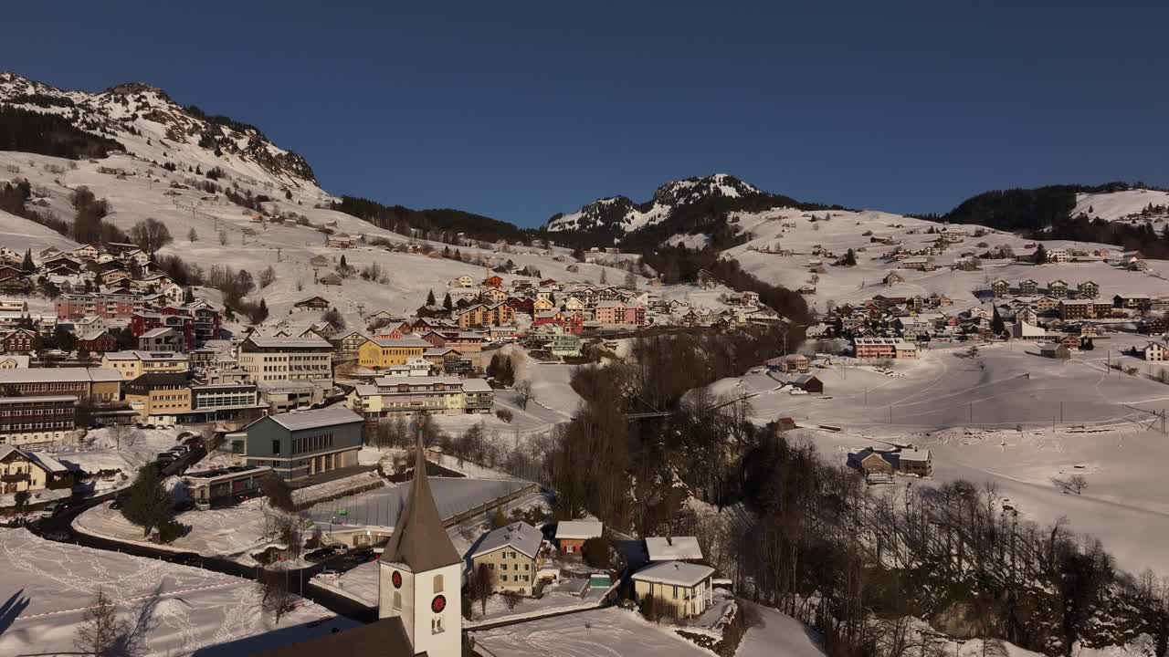 Aerial view of Amden, Switzerland, a picturesque village nestled amidst snow-capped mountains. The iconic St. Gallus Church stands proudly, a testament to the region's rich history.