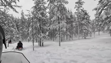 Riding-snowmobiles-through-snowy-woods-in-Finland,-passing-by-cabins-with-snow-covered-trees-and-ground