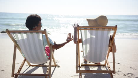 young biracial man and young caucasian woman relax on beach chairs, holding hands