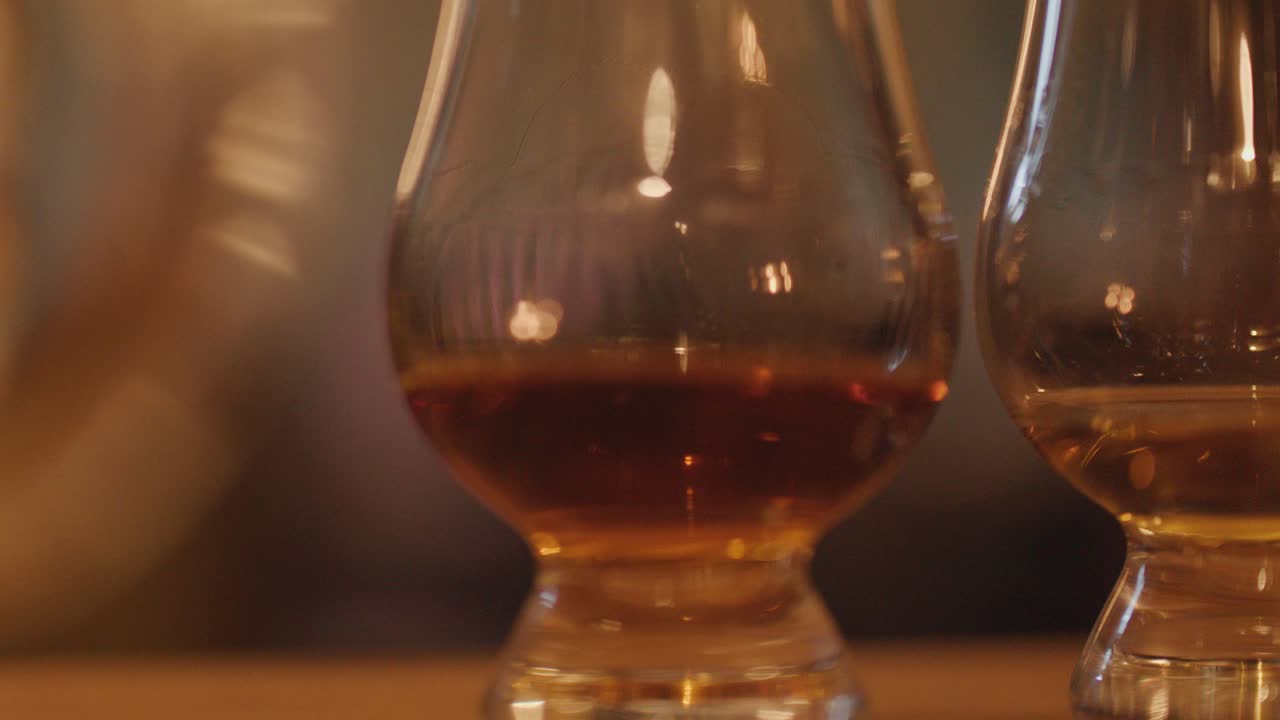 Two whisky glasses with amber liquid sit on a wooden table, softly illuminated by warm candlelight. Gentle camera movement creates a dreamy, intimate atmosphere
