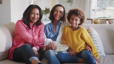 Feliz-Grupo-Familiar-Femenino-De-Tres-Generaciones-Sentado-Viendo-Televisión,-Riendo-Y-Comiendo-Palomitas-De-Maíz-Juntas