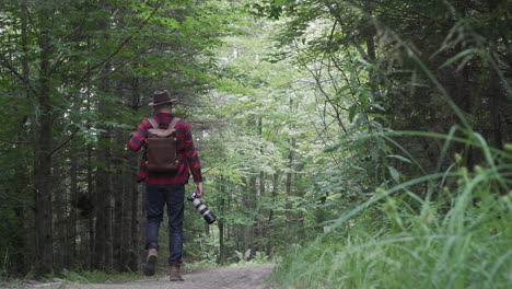 A-young-male-adult-photographer-with-a-large-camera-in-his-hand-walks-down-an-open-forest-road-surrouneded-by-green-fauna-and-trees