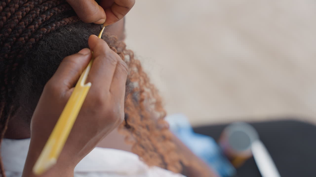 Close-up de estilista negra instalando extensões usando gancho amarelo, mãos laçando cabelo sintético encaracolado na base de trança nagô, tensão controlada, técnica focada, fundo de área de trabalho desfocado, profissional.