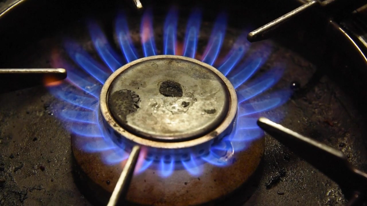 Primo piano di un fornello a gas da cucina che si accende con una vivace fiamma blu. Una padella con olio caldo sfrigola e bolle, pronta per cucinare.