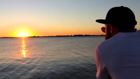 bearded man with cap staring at the sunset on a river deck