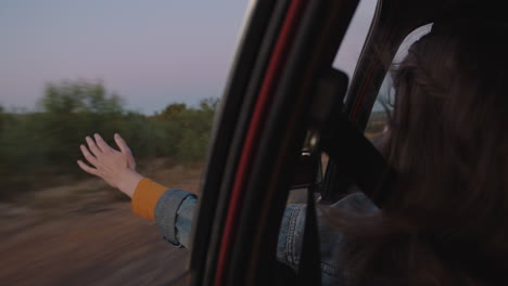 woman in car holding hand out window feeling wind blowing through fingers driving in countryside on road trip travelling for summer vacation enjoying freedom on the road at sunrise