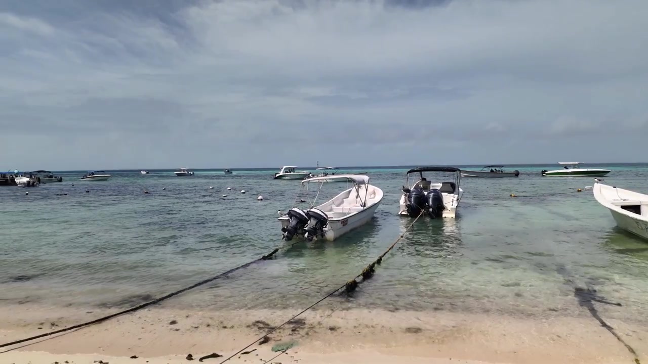 Ripresa panoramica a livello degli occhi con panning a sinistra di diverse piccole imbarcazioni a motore e motoscafi ormeggiati vicino alla riva di una tranquilla spiaggia tropicale. L'acqua cristallina del Mar dei Caraibi rivela le texture sabbiose