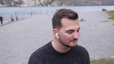 young man sits on the shore of a lake and bounce his head to the rhythm of the music from his earphones - tracking shot