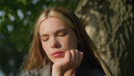 lady in grey clothing sitting under tree with her head resting on her right hand, sunlight softly illuminating her face, she appears deep in thought, eyes slightly closed, as she holds an open book