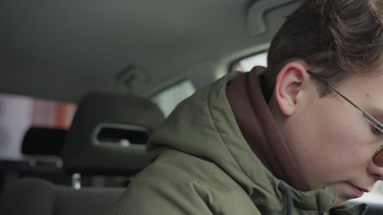 young boy wearing green winter jacket sits inside vehicle, closes car door and adjusts front seat, with soft daylight entering from window and modern residential buildings in blurred background