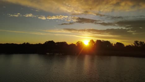 Eine-Dynamische-Luftaufnahme-Eines-Sonnenuntergangs-Mit-Sonnenlicht,-Das-Durch-Die-Bäume-Läuft,-Während-Man-über-Einen-Fluss-In-Der-Argentinischen-Stadt-Córdoba-Fliegt