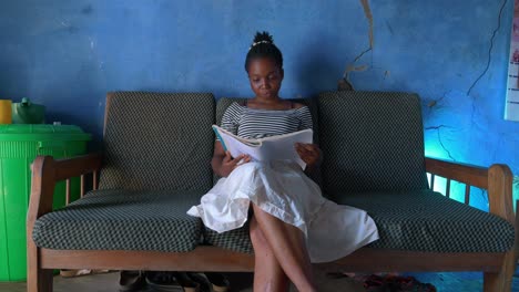 black female young woman from poor village in africa studying reading a book in her humble house while sitting on a sofa couch