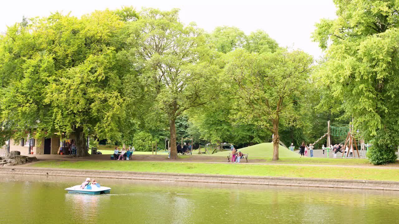 Small paddle boat moves across calm lake in bright, green park with people and trees