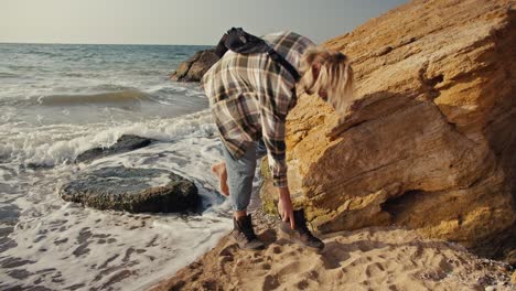 un tipo rubio con una camisa a cuadros salta de una piedra a la playa y trata de ponerse sus zapatos en la orilla rocosa de la playa cerca del mar por la mañana