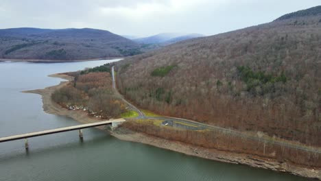 A-drone-flying-high-along-a-mountain-road,-then-panning-sideways-over-a-mountain-lake-and-bridge-during-early-winter-in-the-northern-Appalachian-mountains