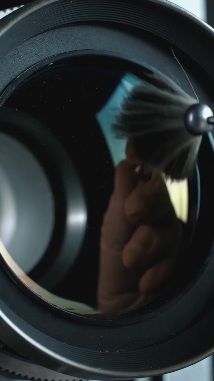 Vertical video: Seeing specks, gloved hand brushing front lens at bench, removing dust with brush