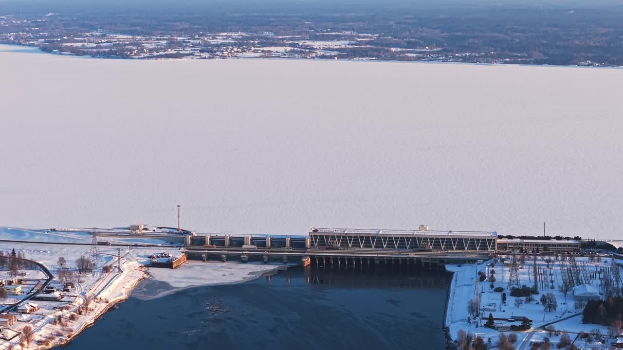 Ampio impianto idroelettrico con fiume ghiacciato e argini coperti di neve, panoramica con teleobiettivo.