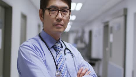 Porträt-Eines-Asiatischen-Männlichen-Arztes-Mit-Verschränkten-Armen-Lächelnd,-Während-Er-Im-Flur-Des-Krankenhauses-Steht
