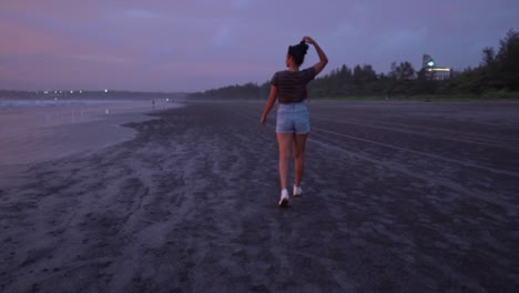 landscape view of a young woman walking on a sandy beach by the ocean, during a purple sunset, while she unite her hair