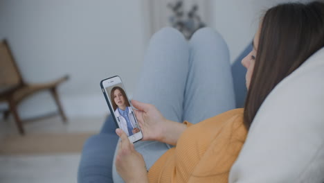 Una-Mujer-Tumbada-En-El-Sofá-Hablando-Con-Una-Doctora-En-Vídeo-Usando-Un-Teléfono-Inteligente.-Consulta-Médica-Remota-Con-Un-Médico-A-Través-Del-Teléfono-Móvil