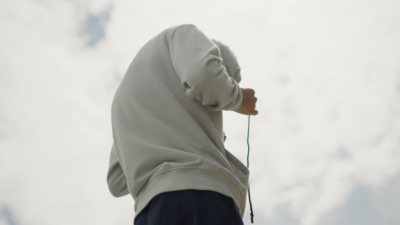 persona en ángulo bajo sujetando una cuerda contra un cielo nublado, preparándose para un movimiento con una silueta dramática y detalles de la sudadera con capucha, manos enrollando el cordel en un parque, la luz nublada enfatiza la postura y el movimiento, ambiente