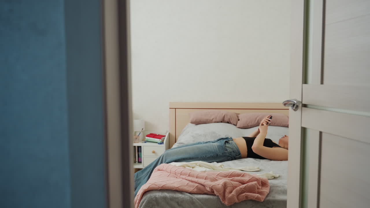White Woman Lying On Bed Scrolling Phone, Quiet Bedroom Perspective From Doorway, Relaxed Recline With Jeans And Earbuds, Modern Minimal Decor And Soft Blanket, Solo Downtime And Reflective Digital