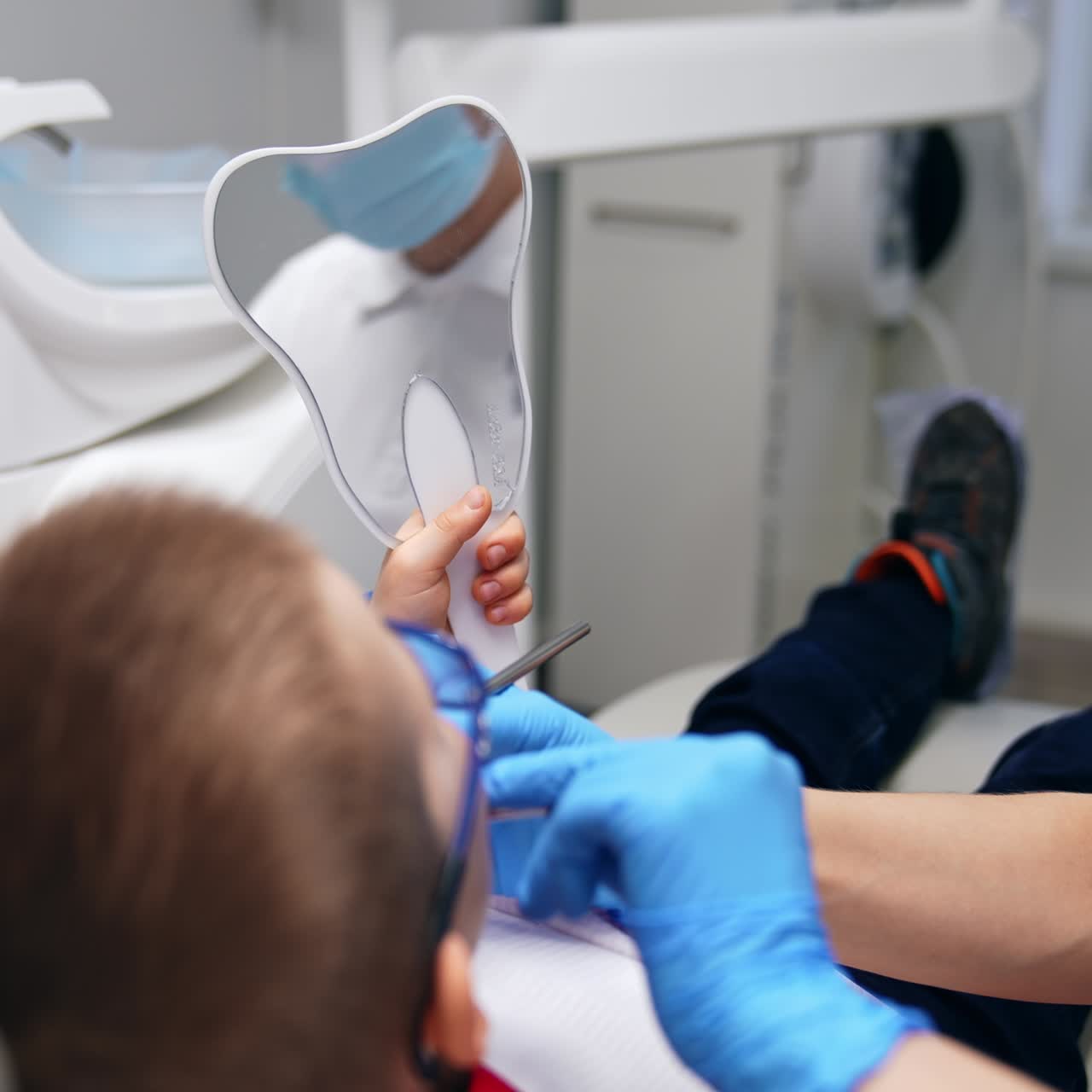 Bambino sorridente che si guarda allo specchio. La dentista mostra il risultato del suo lavoro con l'aiuto di strumenti in metallo