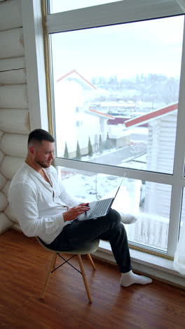 man sitting on the chair near the window holds a laptop on his knee. man works comfortably at home away from office. vertical video.