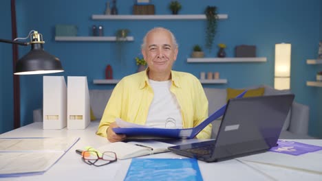 Home-office-worker-old-man-smiling-at-camera-looking-at-paperwork.
