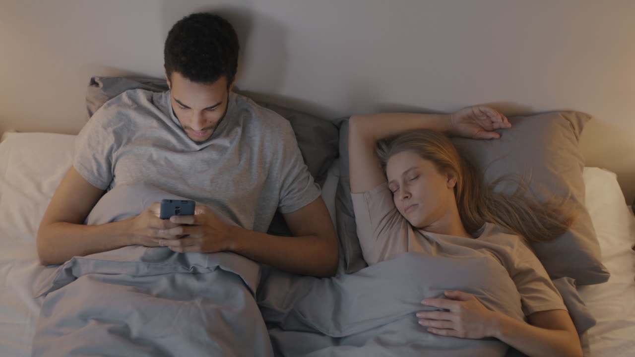Couple in bed, one using a phone while the other sleeps