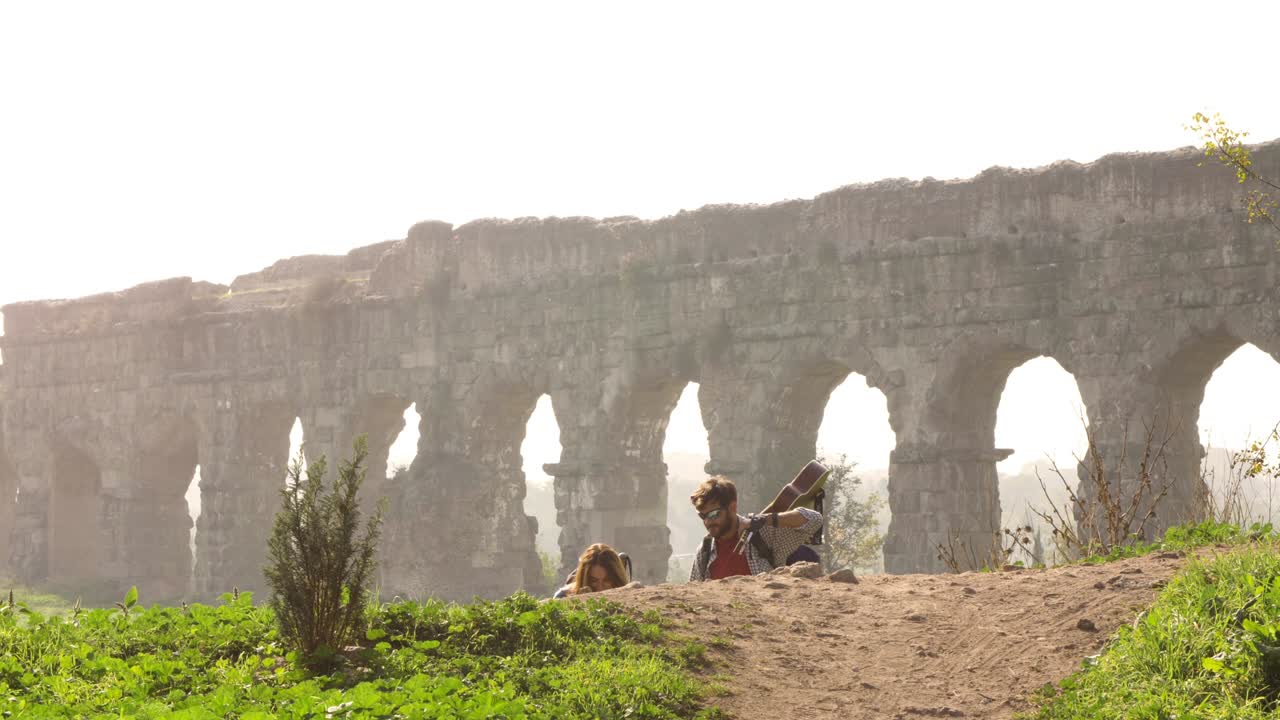 Ungt, nydelig par ryggsekkturister går hånd i hånd mot romerske akveduktbuer i ruinene av Parco degli Acquedotti i Roma ved en romantisk, tåkete soloppgang, med gitar og sovepose. Sakte film.