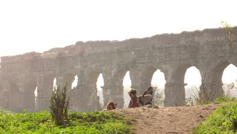 young lovely couple backpackers tourists walking holding hands toward roman aqueduct arches in parco degli acquedotti park ruins in rome on romantic misty sunrise with guitar and sleeping bag slow motion