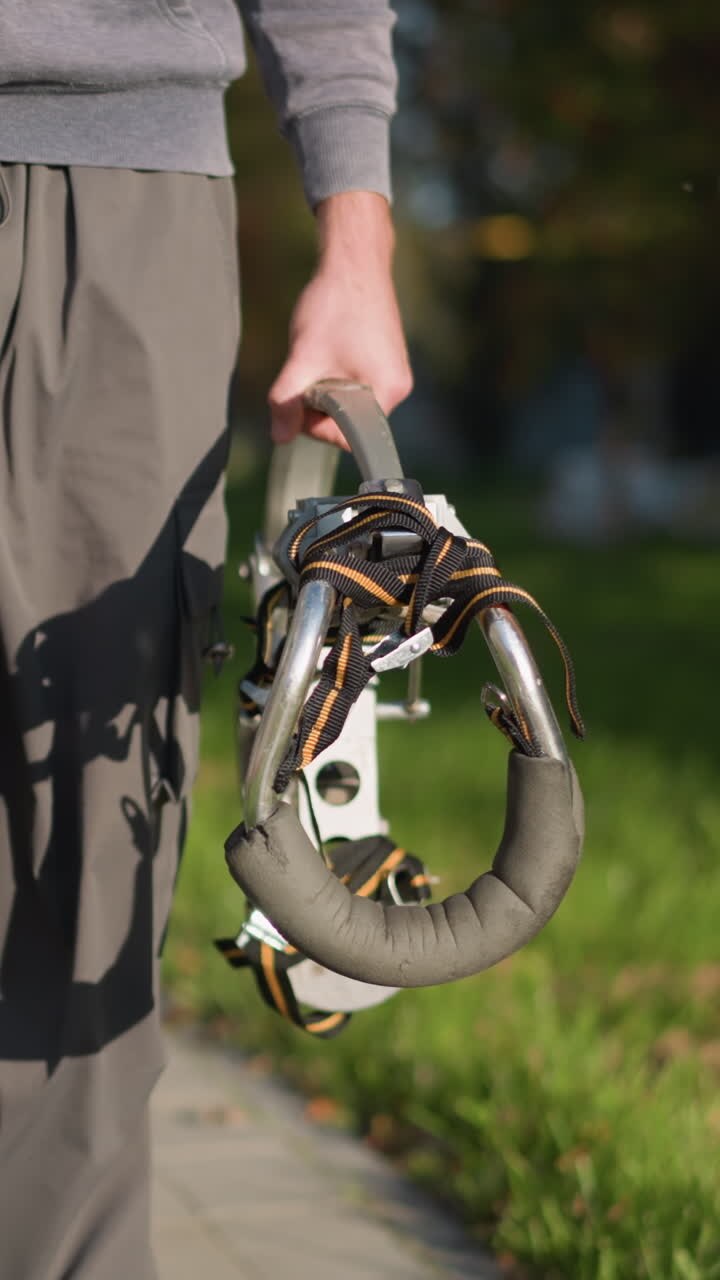 Person in dark gray sweatpants and matching hoodie holding metallic stilts with orange and black straps in blurred grassy outdoor area, focused on lower body, showcasing balance and movement