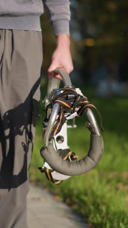 persona con pantalones de deporte gris oscuro y sudadera con capucha a juego sosteniendo pilotes metálicos con correas naranjas y negras en un área al aire libre cubierta de hierba borrosa, centrada en la parte inferior del cuerpo, mostrando equilibrio y movimiento