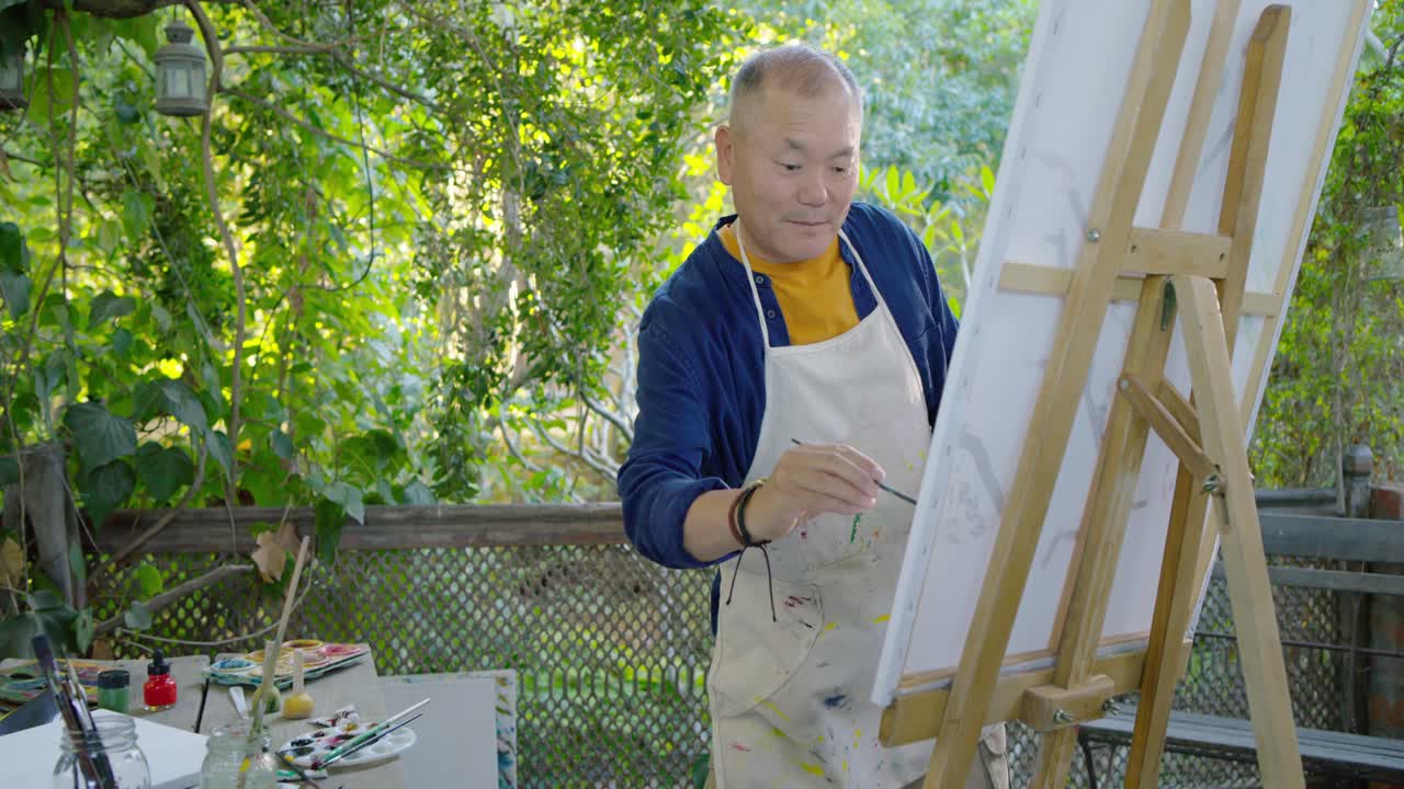 Senior Chinese man wearing apron inspired by garden deck selecting brush mixing refining canvas